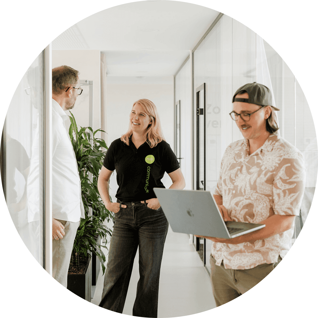Drei Personen stehen und gehen in einem modernen Büroflur mit Glaswänden; eine Person hält ein Laptop, eine andere trägt ein schwarzes Polo mit Firmenlogo, im Hintergrund Pflanzen und offene Türen.