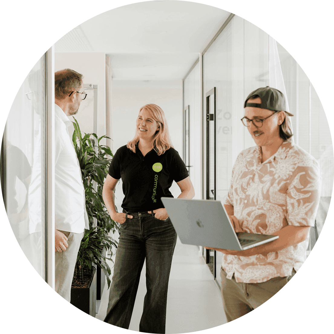Three people are standing and walking in a modern office hallway with glass walls; one person is holding a laptop, another is wearing a black polo shirt with a company logo, and there are plants and open doors in the background.