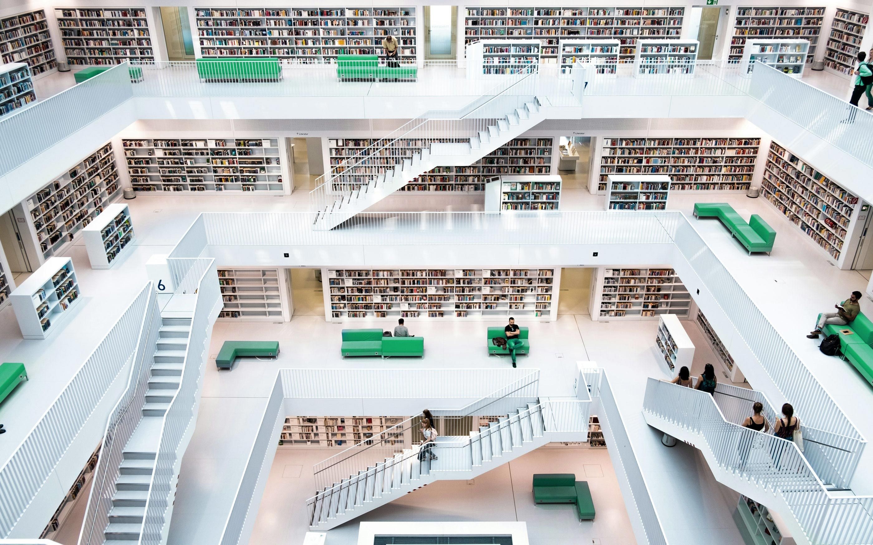 Innenansicht einer großen, modernen Bibliothek mit mehreren offenen Ebenen, weißen Treppen und Geländern. Die Wände sind vollständig mit Bücherregalen gefüllt. Auf den Etagen stehen grüne Sitzmöbel, auf denen einige Menschen verteilt sitzen oder stehen.