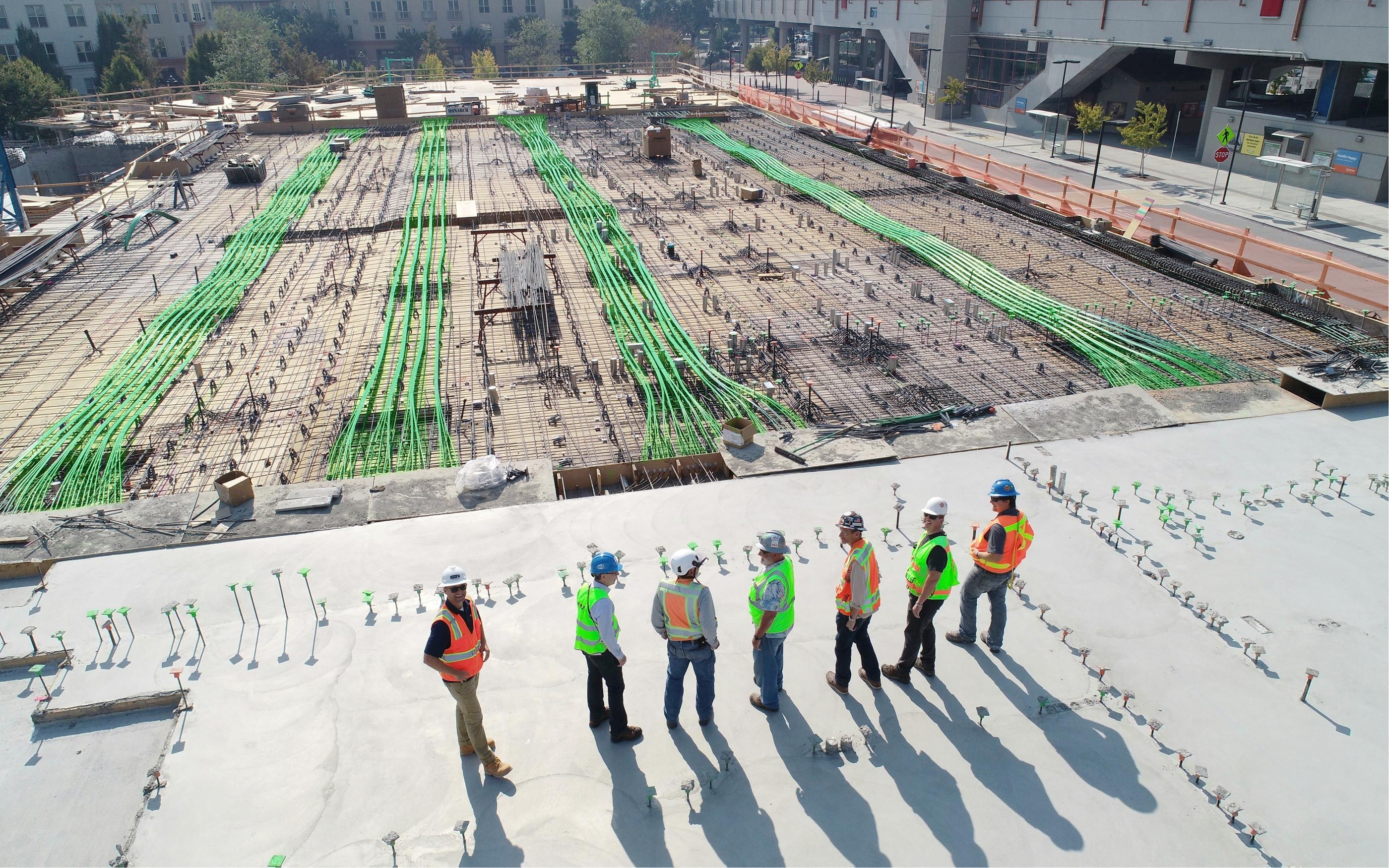 Sechs Personen in Sicherheitswesten und Schutzhelmen stehen auf einer Baustelle zusammen und sprechen miteinander. Im Hintergrund sind große Betonflächen, zahlreiche grüne Installationsleitungen und Baustellenmaterialien zu sehen.
